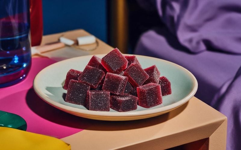 Dark berry cube shaped CBD and CBN sleep gummies in a dish on a nightstand at bedtime